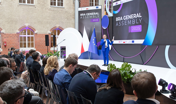 Auf der Bühne der 8ra Konferenz steht eine Frau im blauen Anzug. Im Publikum sitzt eine Vielzahl an Teilnehmenden