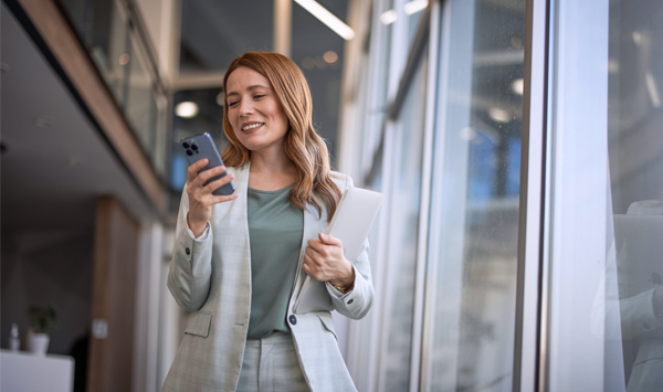 Eine Frau im Blazer läuft durch ein Büro und schaut auf ihr Handy