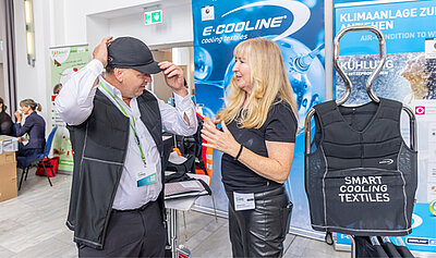 Ein Mann testet an einem Messestand eine kühlende Weste und Mütze aus. Eine Frau berät ihn dabei.