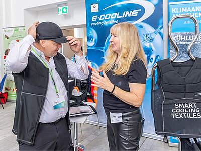 Ein Mann testet an einem Messestand eine kühlende Weste und Mütze aus. Eine Frau berät ihn dabei.
