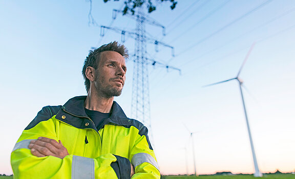 Ein Ingenieur steht auf einem Feld vor einem Strommast und Windrad