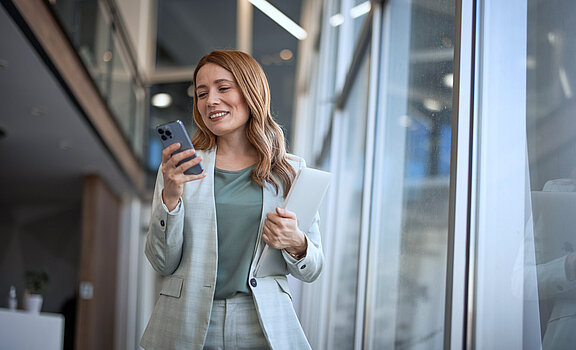 Eine Frau im Blazer läuft durch ein Büro und schaut auf ihr Handy